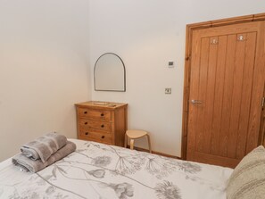 Une chambre à coucher comprenant un lit, une commode en bois, un petit tabouret, une porte en bois et un miroir fixé au mur.