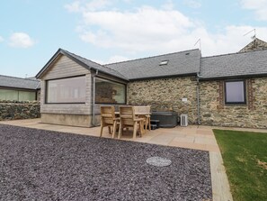 A stone building with a large window, a hot tub, and a patio with wooden chairs. - 2 Trem Y Mor (Holyhead)