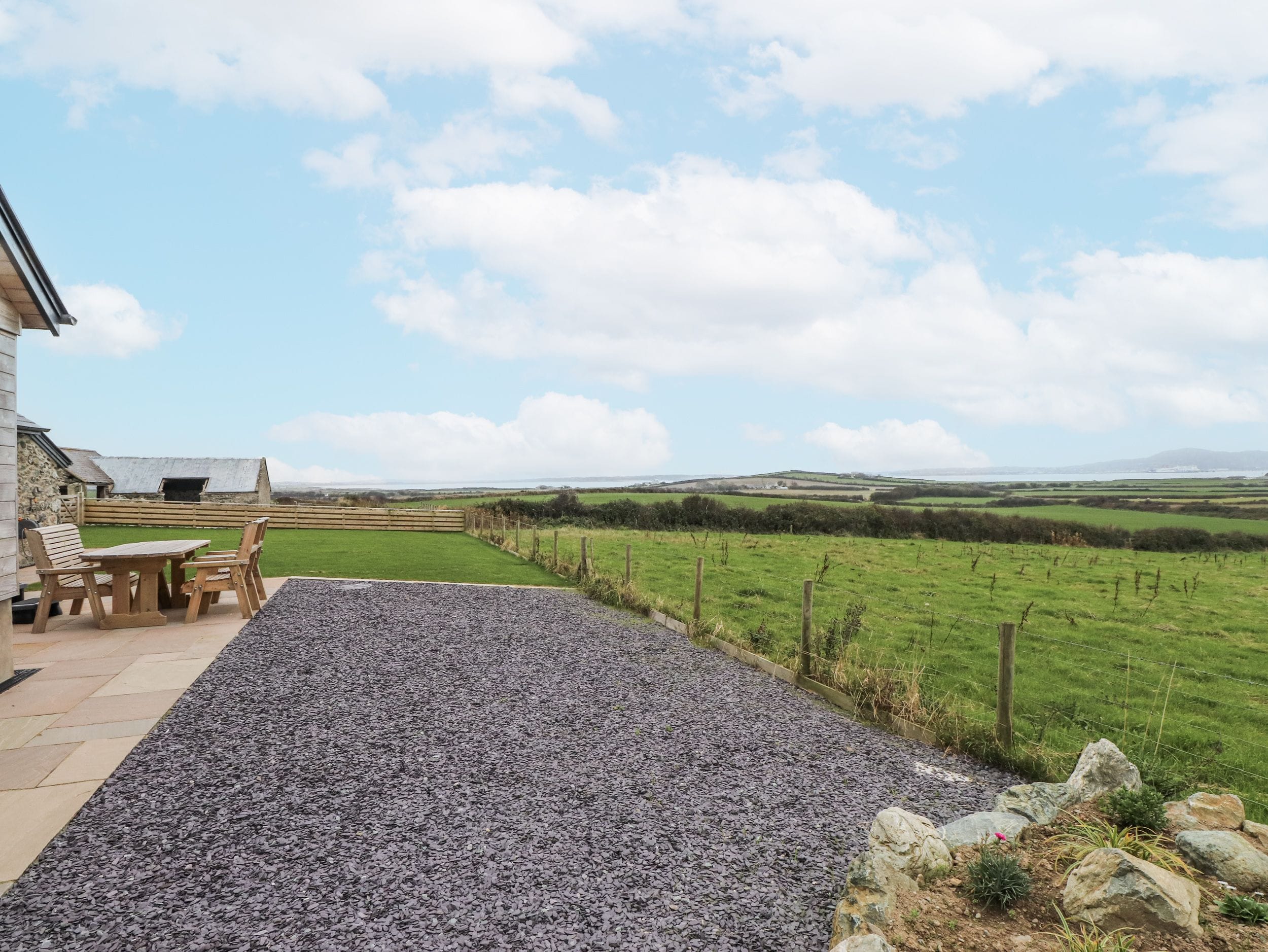 A patio area with a gravel surface, wooden furniture, and a view of a green field and distant hills.