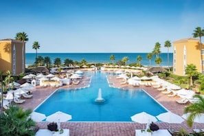 A resort pool with a central fountain, surrounded by lounge chairs and umbrellas, with palm trees and a view of the ocean. - Palacio de Sancti Petri Gran Melia (Novo Sancti Petri)