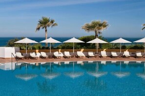 A poolside area with lounge chairs and umbrellas, palm trees, and a clear view of the ocean.