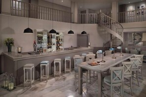 Un área de bar espaciosa con techo alto, una isla central con taburetes y una escalera que lleva a un nivel superior.