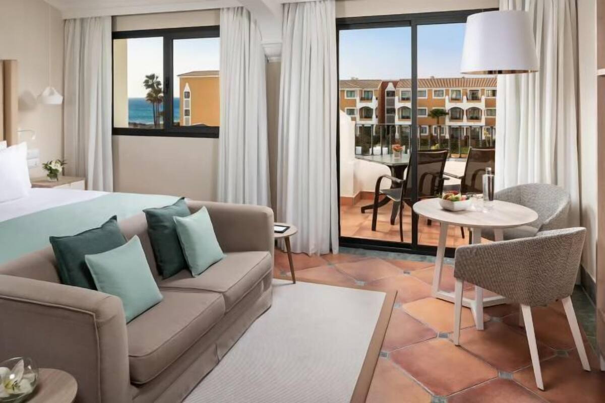 Ein Hotelzimmer mit einem Bett, einem Sofa, einem Esstisch und einem Stuhl. Im Hintergrund ist ein Pool und Gebäude zu sehen.