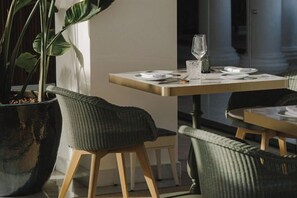 A dining area with a table set for two, a potted plant, and wicker chairs.