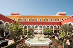 A courtyard with a central fountain, surrounded by trees and red flowers, and a building with arches and a yellow facade. - Palacio de Sancti Petri Gran Melia (Novo Sancti Petri)