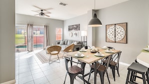 A dining area with a table set for four, chairs, a ceiling fan, and a view of a swimming pool through large windows. - 3BR Encantada Townhome, Heated Pool (Kissimmee)