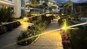 Un chemin mène à un bâtiment moderne doté de balcons, entouré de jardins aménagés et de palmiers.