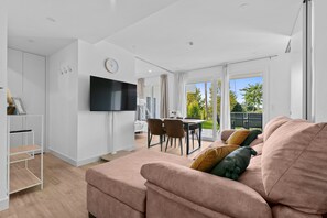 A modern living room with a sofa, a flat-screen TV, a dining area with chairs, and a view of the outdoors.