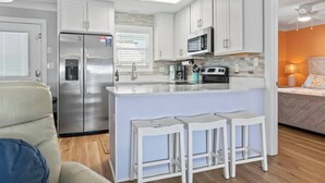 Una cocina moderna con armarios blancos, un refrigerador de acero inoxidable, un microondas y una estufa. Incluye una barra con taburetes y un ventanal con persianas.