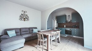 A modern living room with a grey sofa, wooden dining table, and chairs. A kitchen is visible through an archway, featuring dark cabinets and a stainless steel oven. - Casa da Victoria - Apartment 1 (Castrezzato)