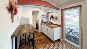 A compact kitchen with a wooden table and bar stools, white cabinets, and a view through a large window. - Charming Apartment with River Views & Sunny Terraces near Lisbon (Vila Franca de Xira)