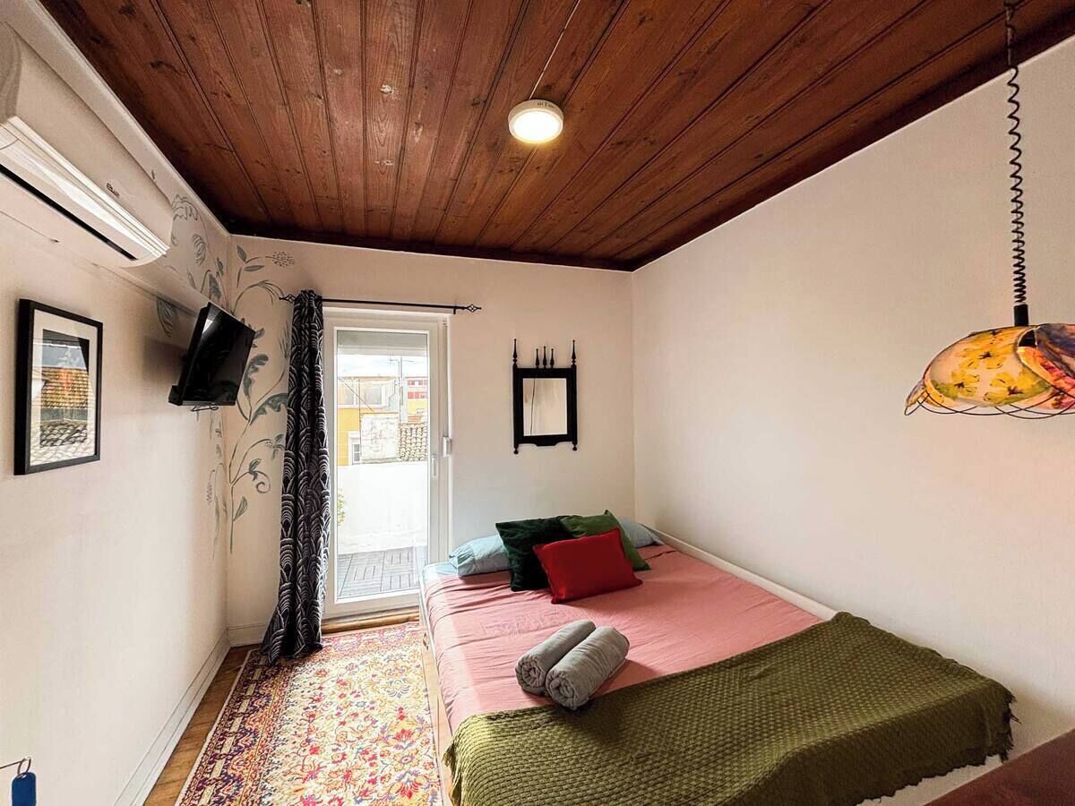 A bedroom with a wooden ceiling, a bed with pink bedding, a green throw, a hanging lamp, and a window with a view of the outside.