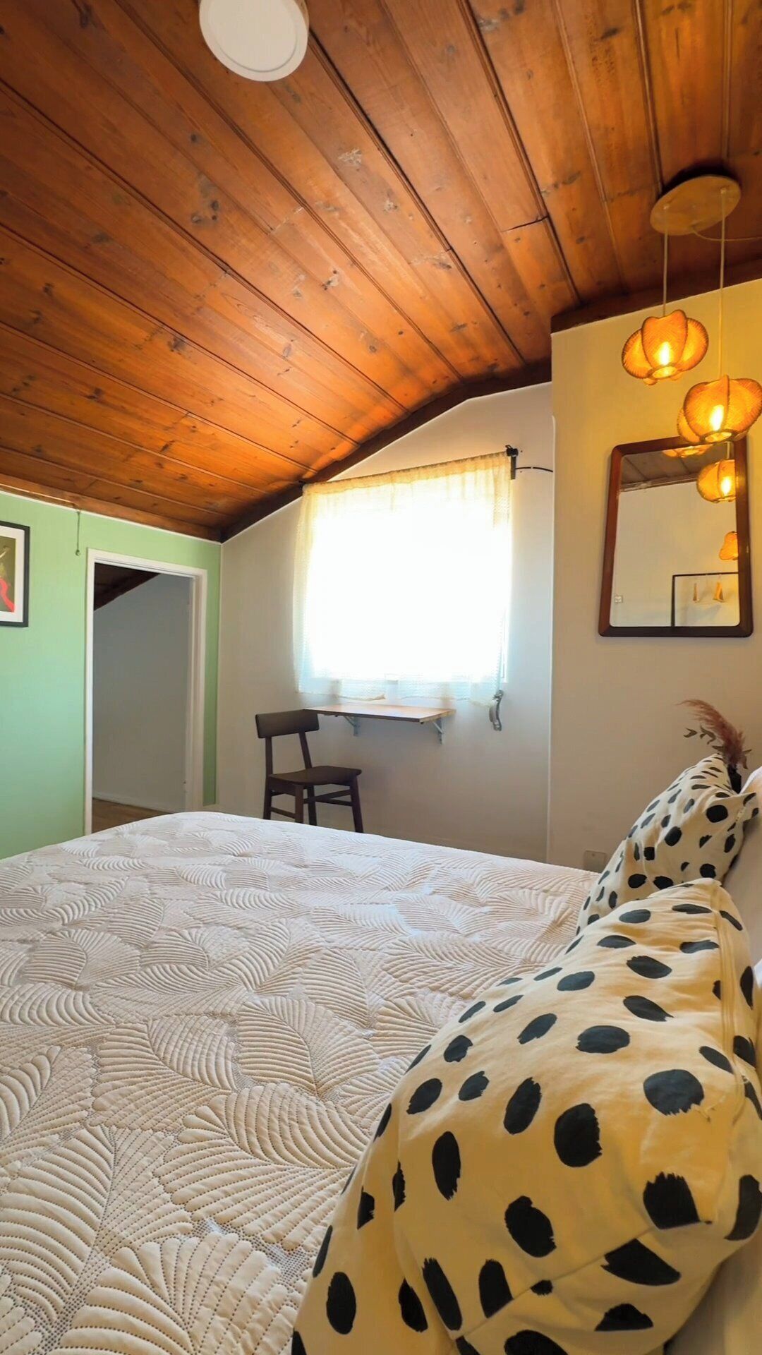 A bedroom with a wooden ceiling, a bed with a patterned quilt, a polka-dotted pillow, a desk with a chair, and a window with curtains.