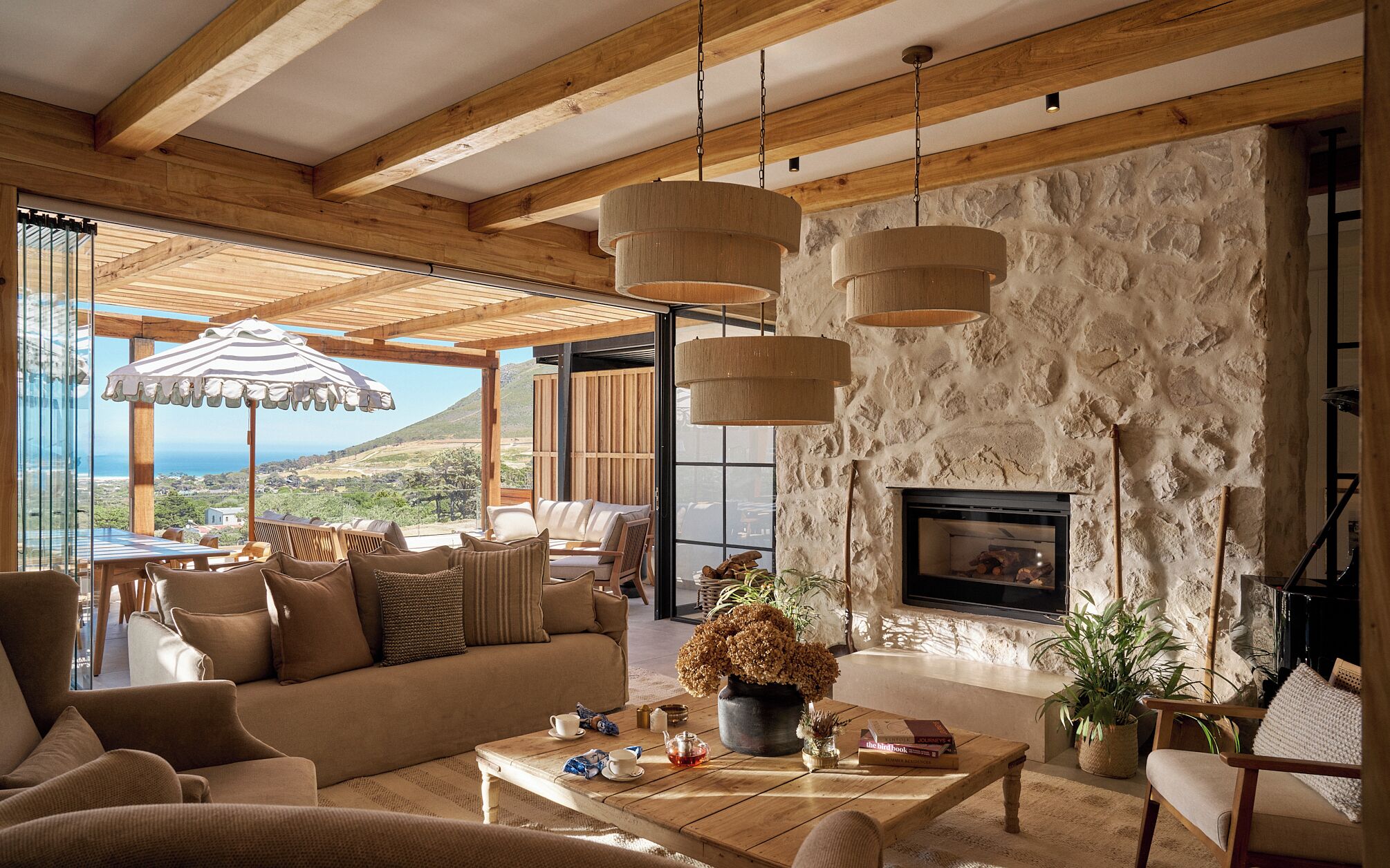 A spacious living room with a stone wall, wooden ceiling beams, and a large window overlooking a scenic landscape.