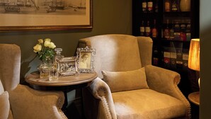 A room with a large armchair, a small round table with a vase of flowers, a framed picture on the wall, and a cabinet with bottles. - The Alexander Estate (Cape Town)