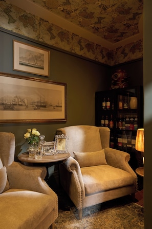 A room with a large armchair, a small round table with a vase of flowers, a framed picture on the wall, and a cabinet with bottles. - The Alexander Estate (Cape Town)