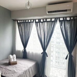 A bedroom with a bed, a window with curtains, an air conditioner, and a basket of towels.