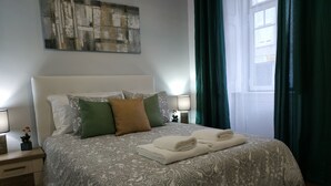 Un lit bien fait, recouvert d’une couvre-lit à motifs, avec deux oreillers et des serviettes pliées. On aperçoit une table de chevet avec une lampe et une petite plante. Un tableau est accroché au-dessus du lit, et des rideaux sont visibles à l’arrière-plan.