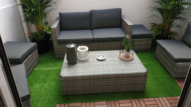 Un petit espace extérieur aménagé avec un canapé gris, deux fauteuils, une table basse en osier et des plantes en pot.
