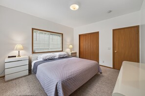 Un dormitorio con cama, dos mesitas de noche con lámparas, una ventana con persianas, un armario de madera y suelo alfombrado.