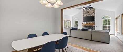 A modern dining area with a white table and blue chairs, a stone fireplace with a TV, and a view of a garden through large windows.