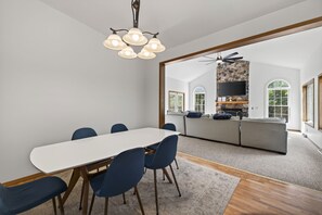 Un comedor moderno con una mesa blanca y sillas azules, una chimenea de piedra con televisor, y vistas a un jardín a través de ventanas amplias.