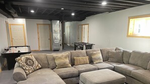 Een woonkamer met een hoekbank, poufs, een tafelvoetbalspel en een groot raam.