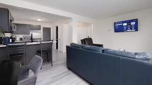 Un salon moderne avec un canapé gris, un coin repas avec une table et des chaises, et une cuisine en arrière-plan.