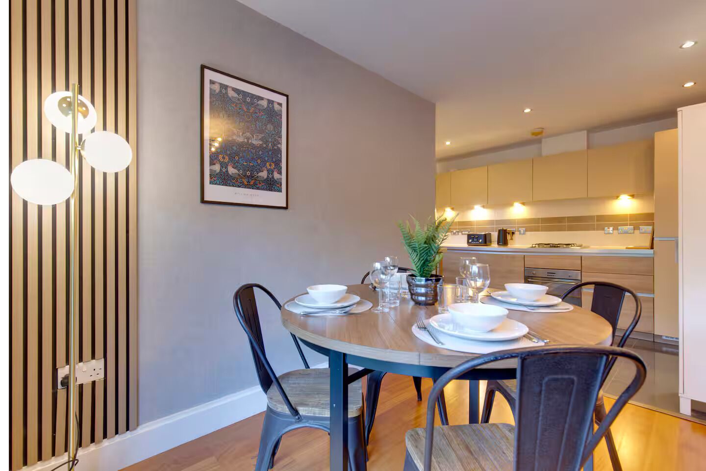 Un coin repas avec une table ronde dressée pour quatre personnes, entourée de chaises. Une œuvre d’art encadrée est accrochée au mur au-dessus de la table.