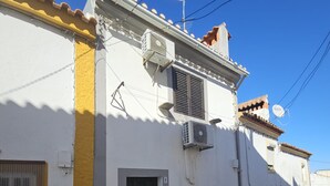 Un bâtiment à la façade blanche, avec une partie jaune sur le côté gauche et un toit recouvert de tuiles. On peut y voir deux unités de climatisation fixées au mur, une antenne parabolique sur le toit et une rue pavée devant.