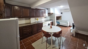 Une cuisine de taille réduite, dotée de meubles de rangement en bois foncé, d’un plan de travail blanc, d’un évier, d’une cuisinière et d’un coin repas avec une table et des chaises.