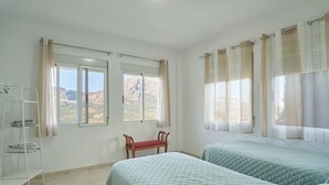 Une chambre avec deux lits, une chaise, une fenêtre avec des rideaux et une étagère où sont pliées des serviettes.