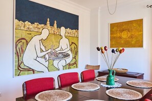 A dining area with a table set for four, red chairs, a large painting, and a vase with fruit on the table.
