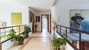Un couloir doté d’un sol carrelé, de plantes en pot, d’œuvres d’art accrochées aux murs et d’un escalier avec une rampe en bois.