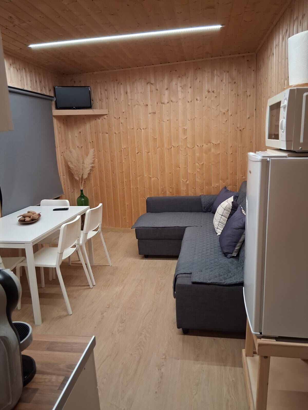 Un espace de vie moderne et compact, avec un intérieur en bois, un canapé gris, une table à manger blanche avec des chaises, une petite télévision et un four à micro-ondes.