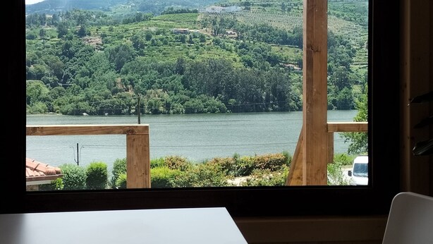 Blick aus einem Fenster mit Holzrahmen auf einen See und grüne Hügel mit verstreuten Gebäuden.