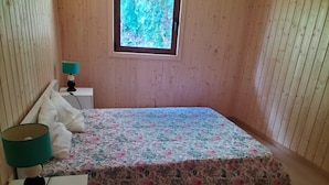 Ein kleines, aufgeräumtes Schlafzimmer mit floralem Bettbezug, einer Nachttischlampe und einem Fenster mit Blick auf Grün.