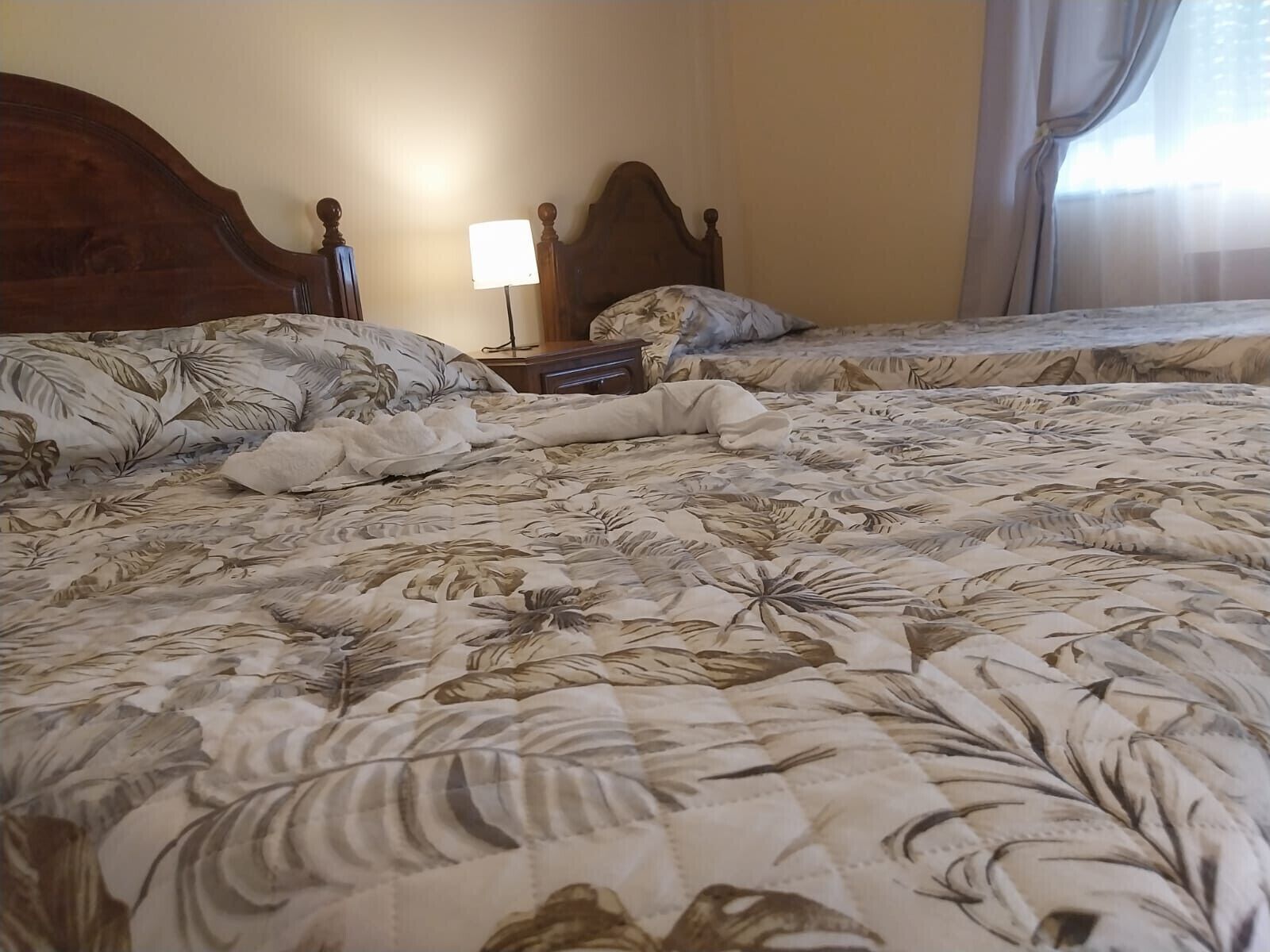 A hotel room with two beds, a nightstand, a lamp, and a window with curtains.