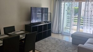 Ett vardagsrum med en TV, ett matbord, stolar, en soffa och en balkong med gardiner.