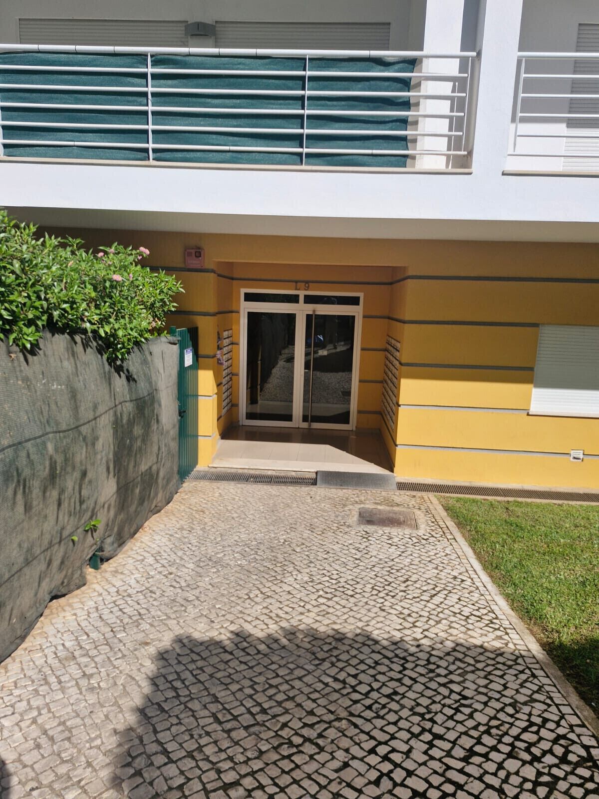 Une entrée d’immeuble avec un chemin dallé menant à une porte en verre, un mur en béton agrémenté de verdure et une façade jaune et blanche.