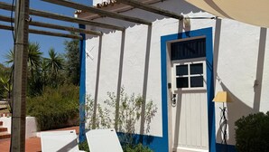 A patio with two white lounge chairs, a white building with blue trim, a door, and a small garden area. - Casa Bungavilia - Roots of Alentejo (Alvito)
