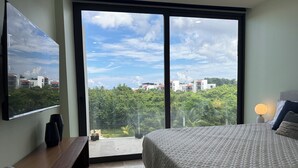 A bedroom with a large bed, a TV mounted on the wall, and a view of a residential area through a sliding door. - Newly Luxury Condo in Cozumel! D'Maria Condos (Cozumel)