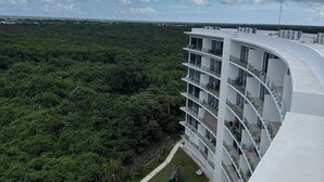 A modern hotel with a curved white facade, multiple balconies, and a lush green garden area. - Newly Luxury Condo in Cozumel! D'Maria Condos (Cozumel)