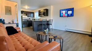 Una sala de estar moderna con un sofá de cuero, un comedor con una mesa pequeña y sillas, una cocina con armarios oscuros y un refrigerador, y un televisor de pantalla plana montado en la pared.