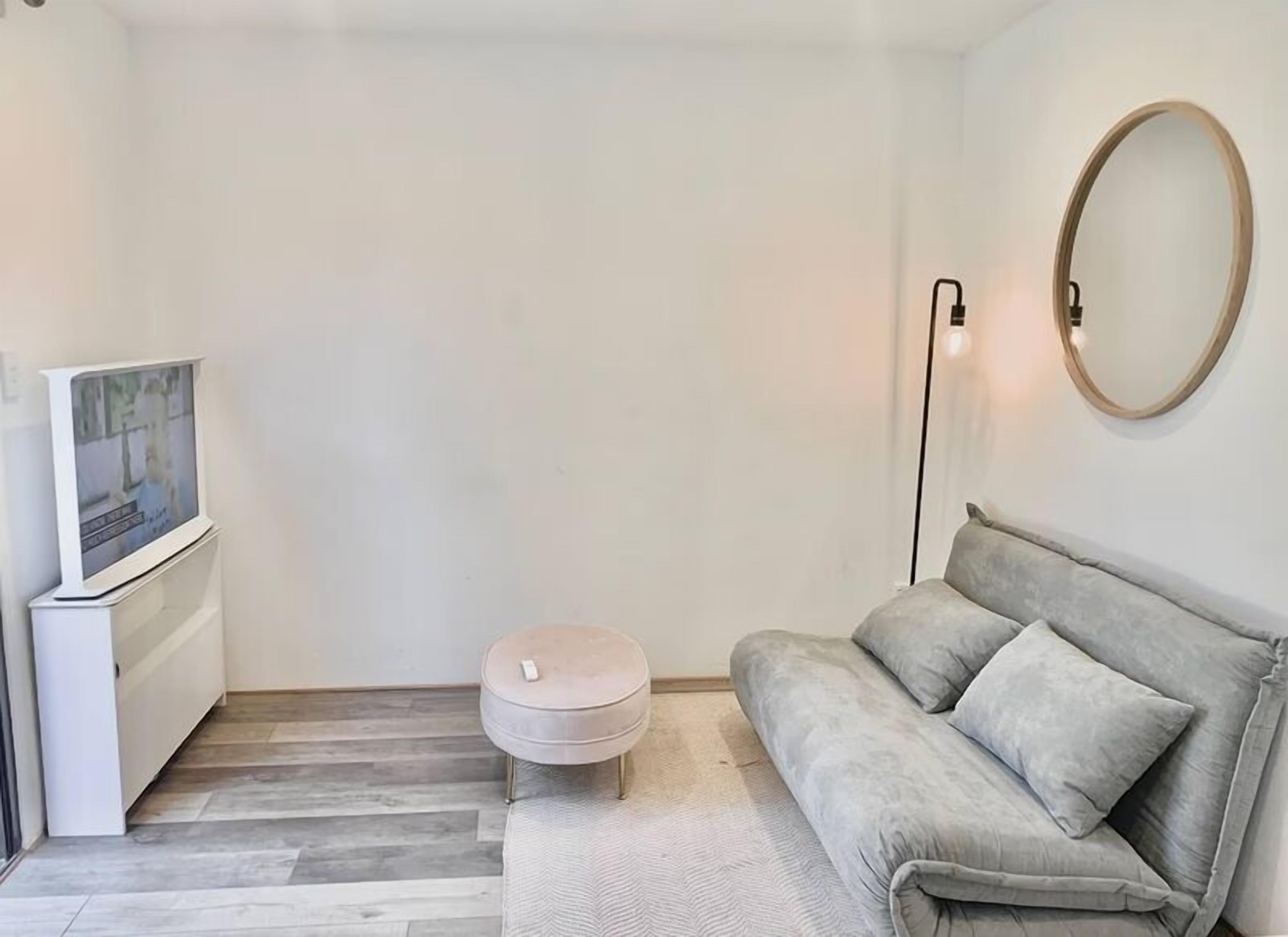 Una sala moderna con un sofá gris, un reposapiés redondo pequeño, un televisor de pantalla plana y un espejo redondo en la pared.