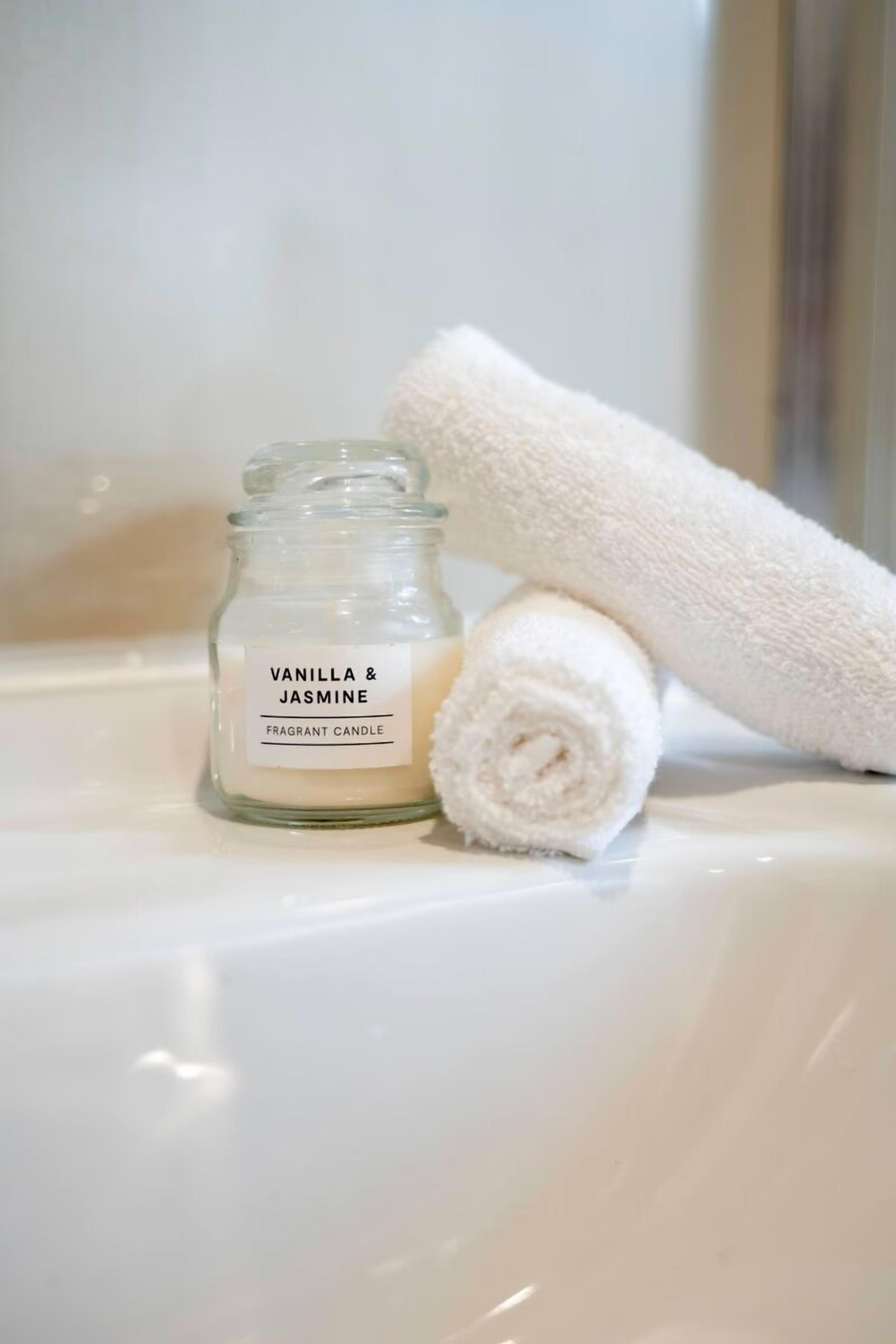 A white scented candle labeled 'Vanilla & Jasmine' and a rolled white towel on a white surface.