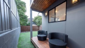 A patio area with two wicker chairs, a wooden deck, and a grey wall with a window. - PP Garden (Bondi)