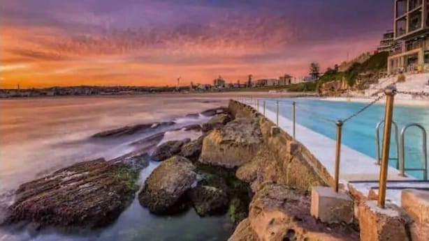 A coastal scene with a rocky shoreline, a swimming pool, and a building with balconies overlooking the water. - PP Garden (Bondi)