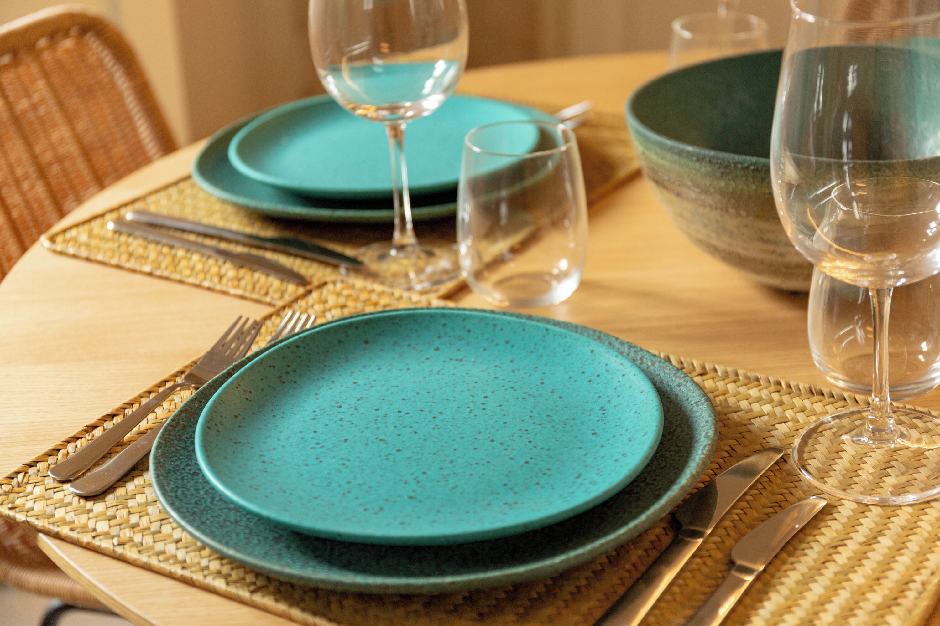 A dining table set with turquoise plates, clear glasses, and silverware, placed on woven placemats.