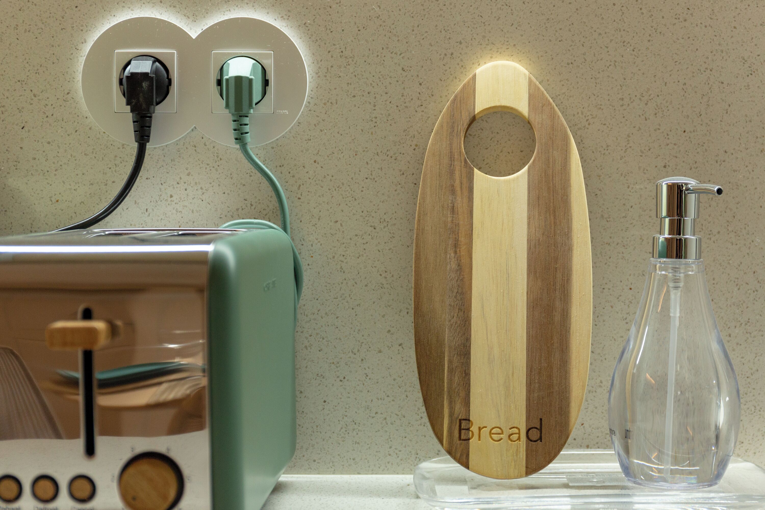 Un tagliere di legno con la scritta 'Bread' incisa, una bottiglia di vetro trasparente con dispenser a pompa e un tostapane con cavo di alimentazione inserito.